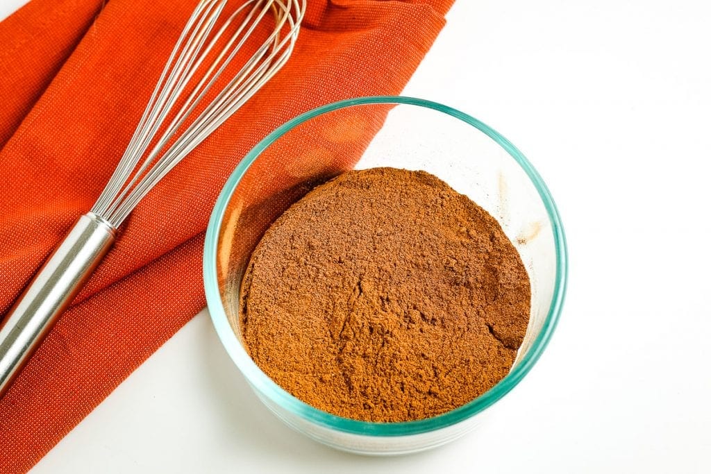 Homemade Pumpkin Pie Spice mixed together in small glass bowl with orange napkin and whisk behind it.