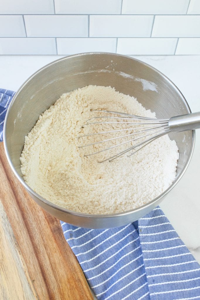 Metal mixing bowl with whisk sitting in it. A mixture of flour, sugar, and salt mixed together.