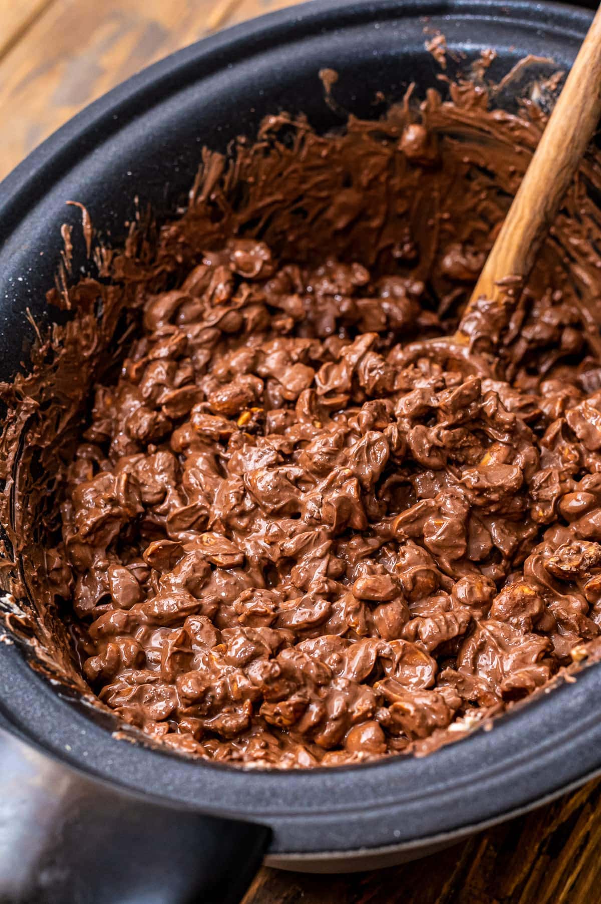 Crock Pot Peanut Clusters mixture in crock pot with wooden spoon sitting in it.