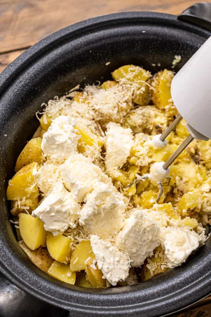 Hand mixer mixing together cooked potatoes, cream cheese, milk in crock pot