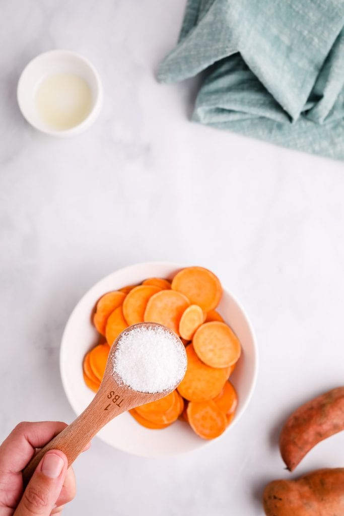 Salt in a teaspoon being held over a bowl of sliced sweet potatoes