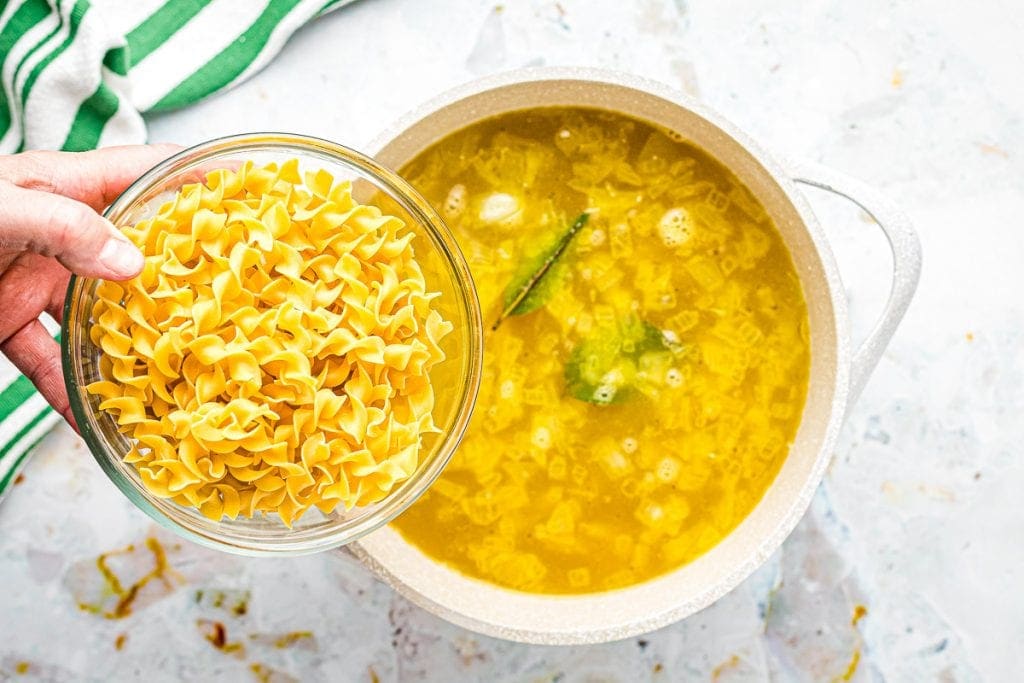 Egg Noodles in glass bowl being added to a pot of chicken noodle soup.