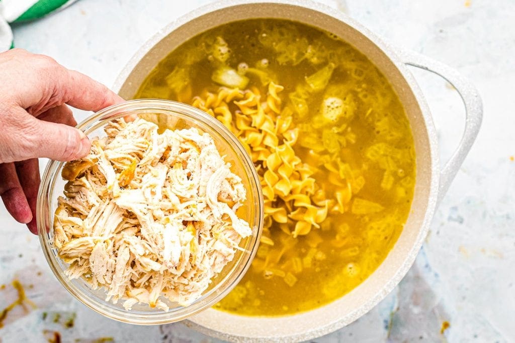 Shredded chicken in glass bowl being added to a pot of chicken noodle soup.