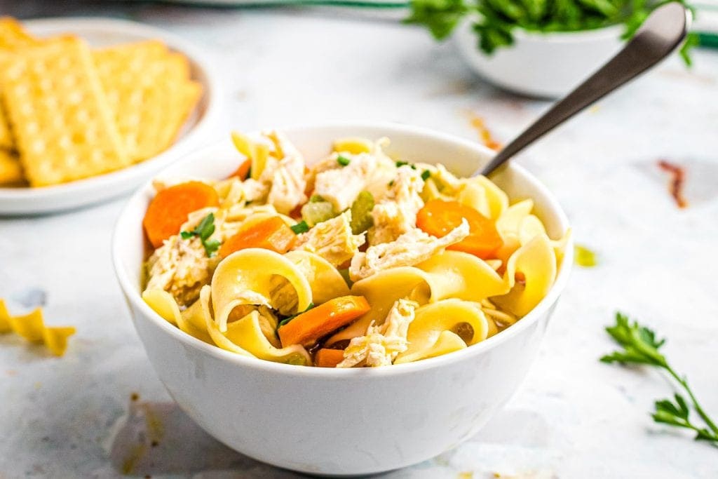 White bowl of Chicken Noodle Soup with soup in it. Crackers on white plate behind it.