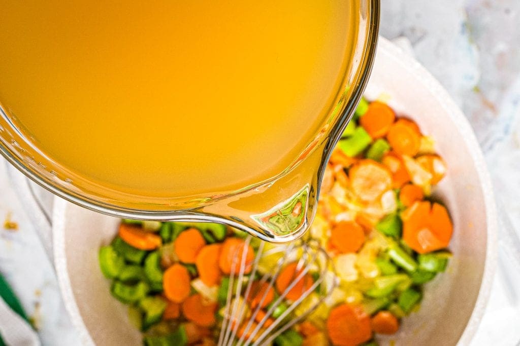 Glass bowl of chicken broth being added to a pot of chicken noodle soup.