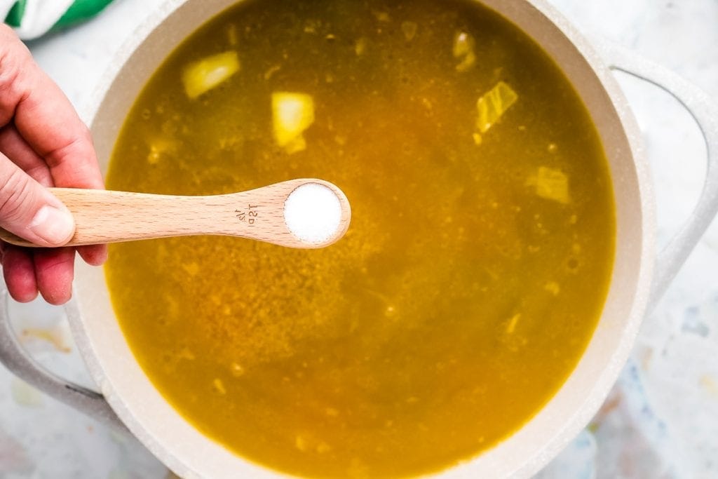 Hand holding spoon of salt and adding it to a pot of chicken noodle soup.