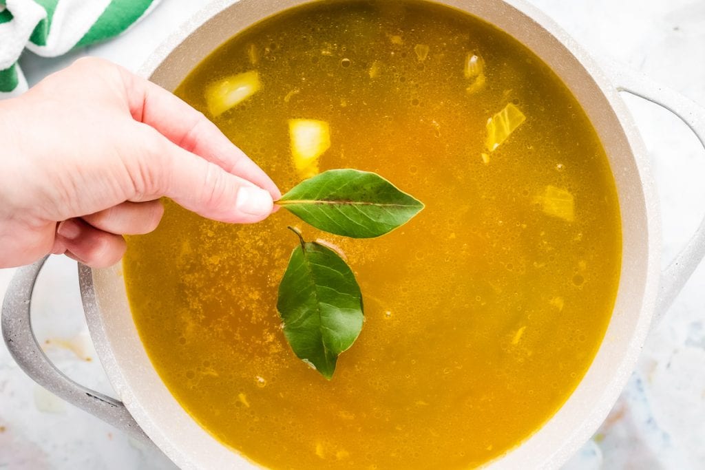 Hand holding bay leaves and adding them to a pot of chicken noodle soup.