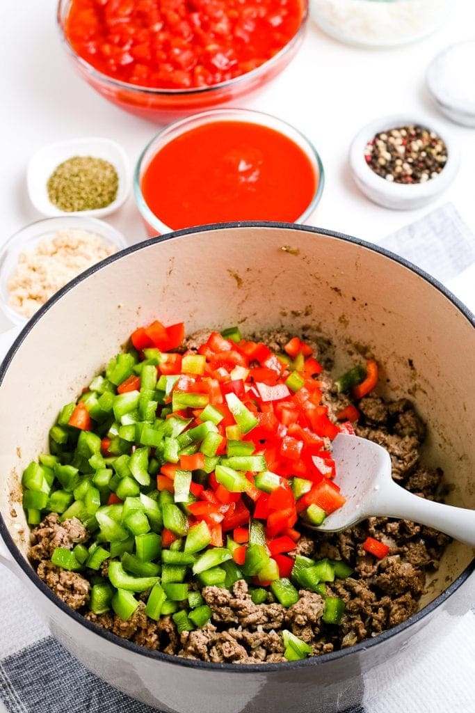 Dutch oven with browned ground beef in it and chopped green and red peppers on top of it.