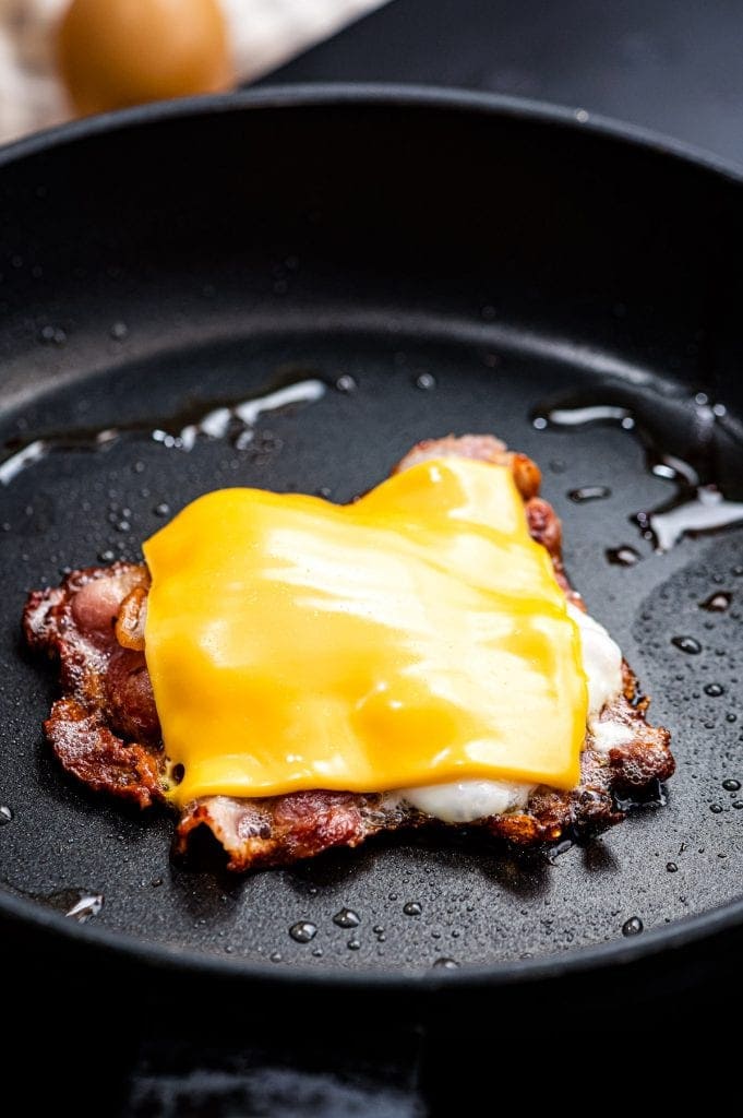 Frying pan with bacon making a shape of a square and a fried egg in the center and melted cheese slice on top.