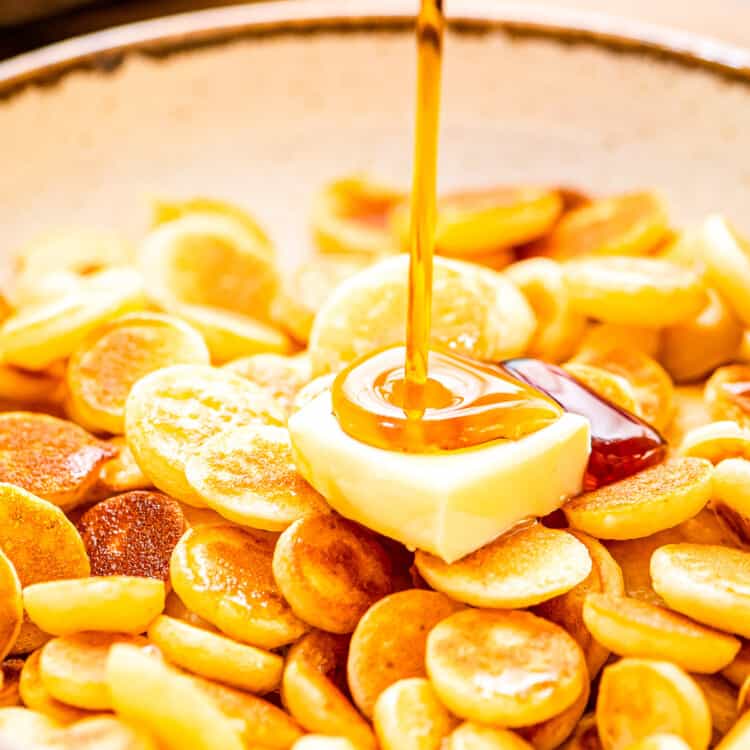 A bowl of mini pancake cereal with a slab of butter and syrup being poured on