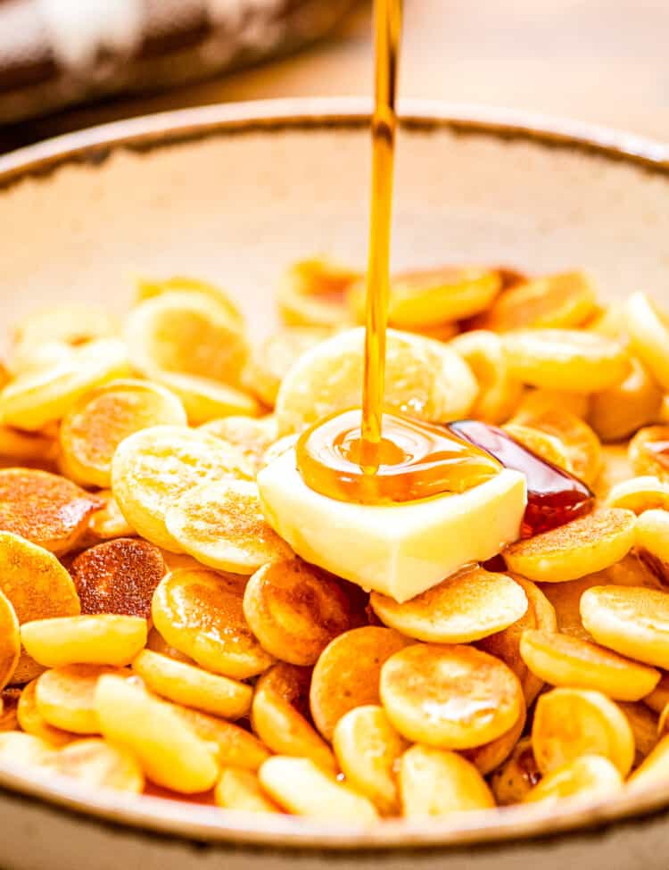 A bowl of mini pancake cereal with a slab of butter and syrup being poured on
