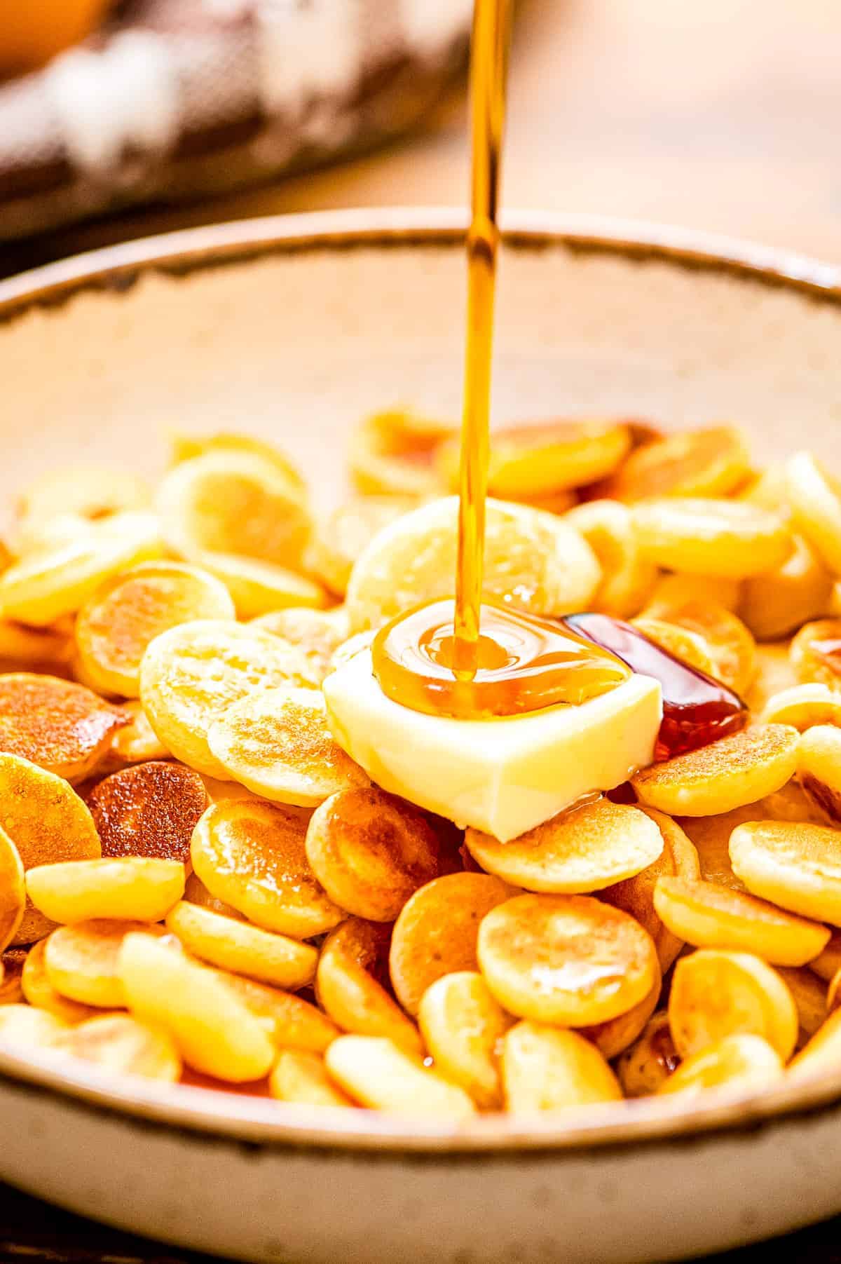 A bowl of mini pancake cereal with a slab of butter and syrup being poured on