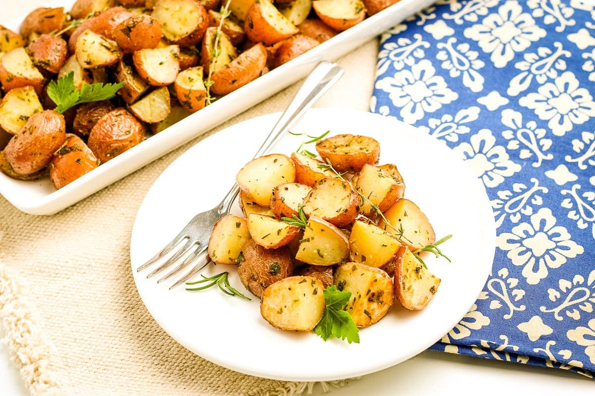 Oven Roasted Potatoes on white plate with fork on it. A serving platter of more potatoes in background and blue napkin.