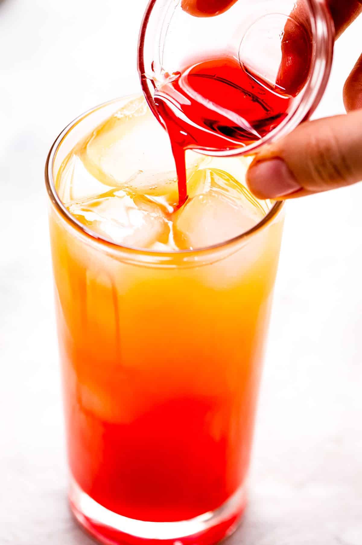 Hand pouring maraschino cherry juice into a high ball glass with orange juice