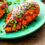 A close-up of grilled BBQ chicken breast with a caramelized, slightly charred glaze, garnished with freshly chopped herbs. The chicken is served on a vibrant turquoise plate, with another piece partially visible in the background.