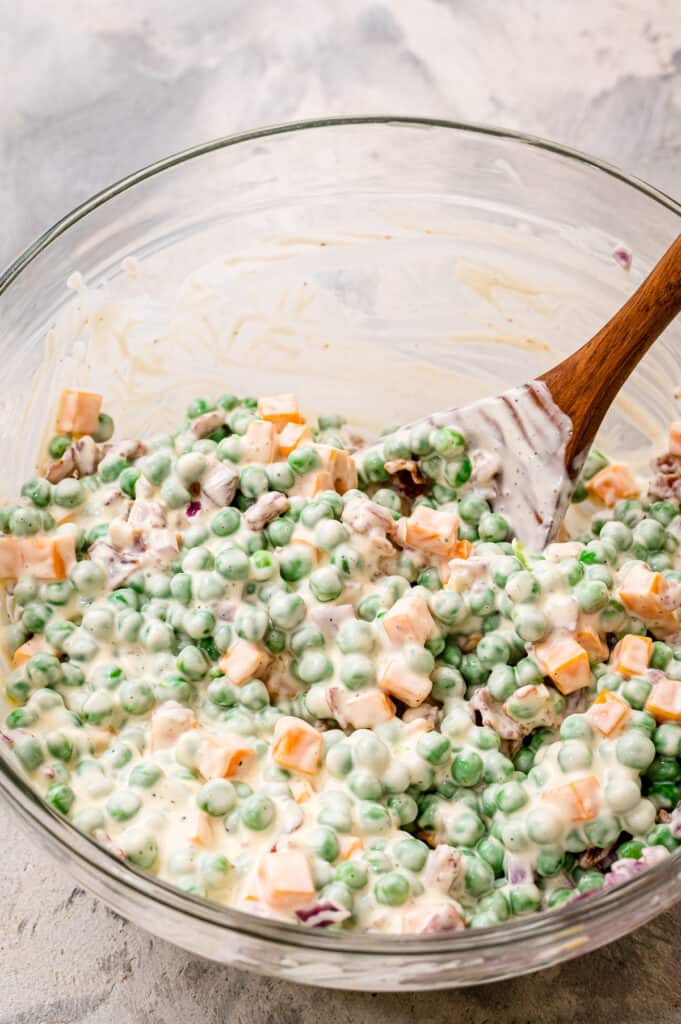 Glass bowl with ingredients for pea salad mixed together with wooden spoon