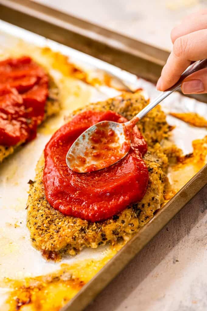 Spoon spreading marinara sauce over breaded chicken breast