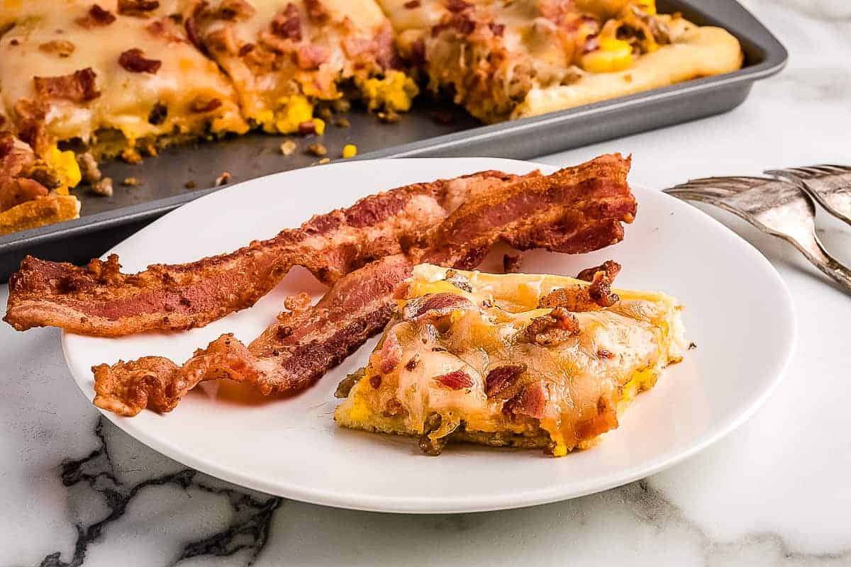 White plate with slice of breakfast pizza and bacon slices