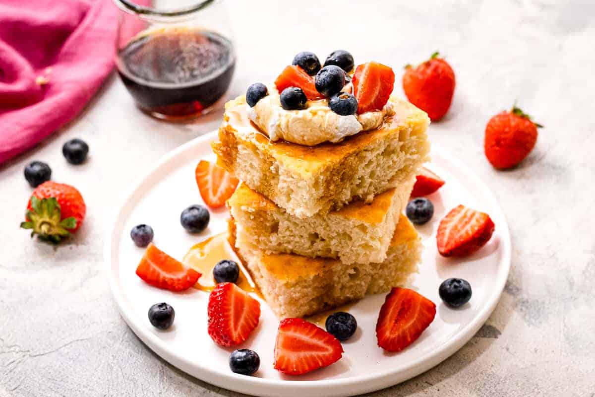 White plate with sheet pan pancakes on it topped with berries and whipped topping