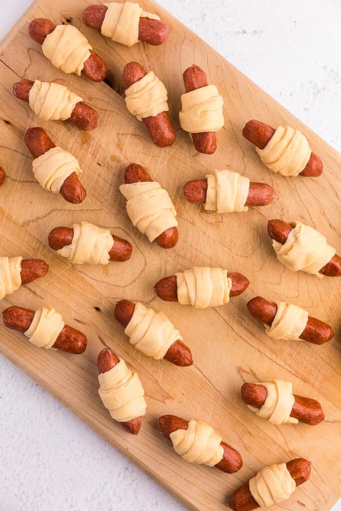 Brown wood cutting board with raw mini pigs in a blanket on it