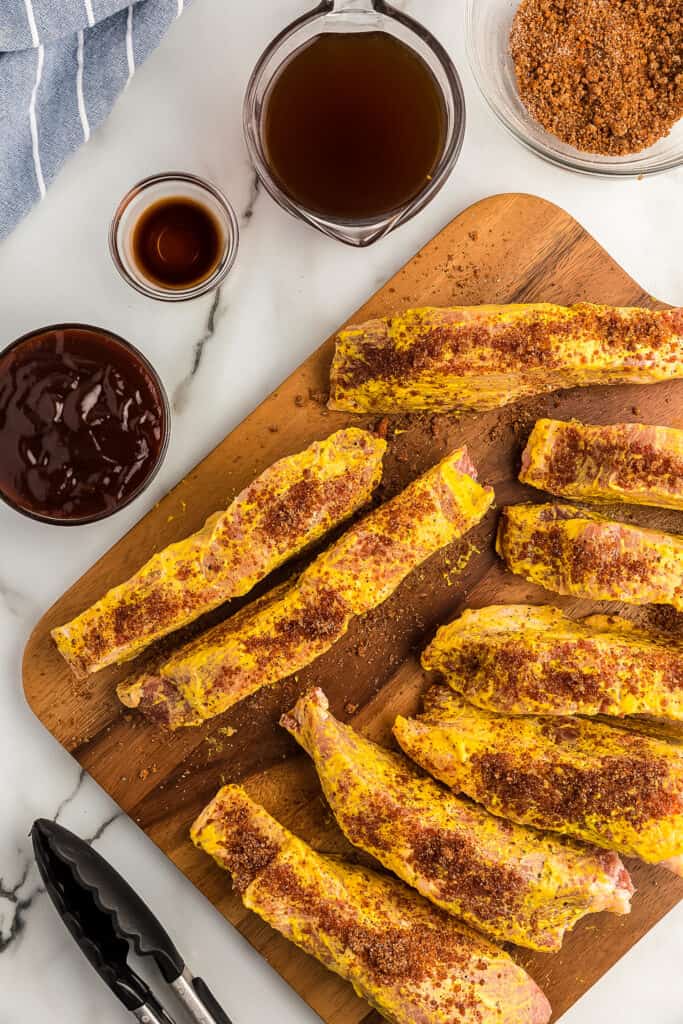 Brown cutting board with ribs covered in spices and yellow mustard