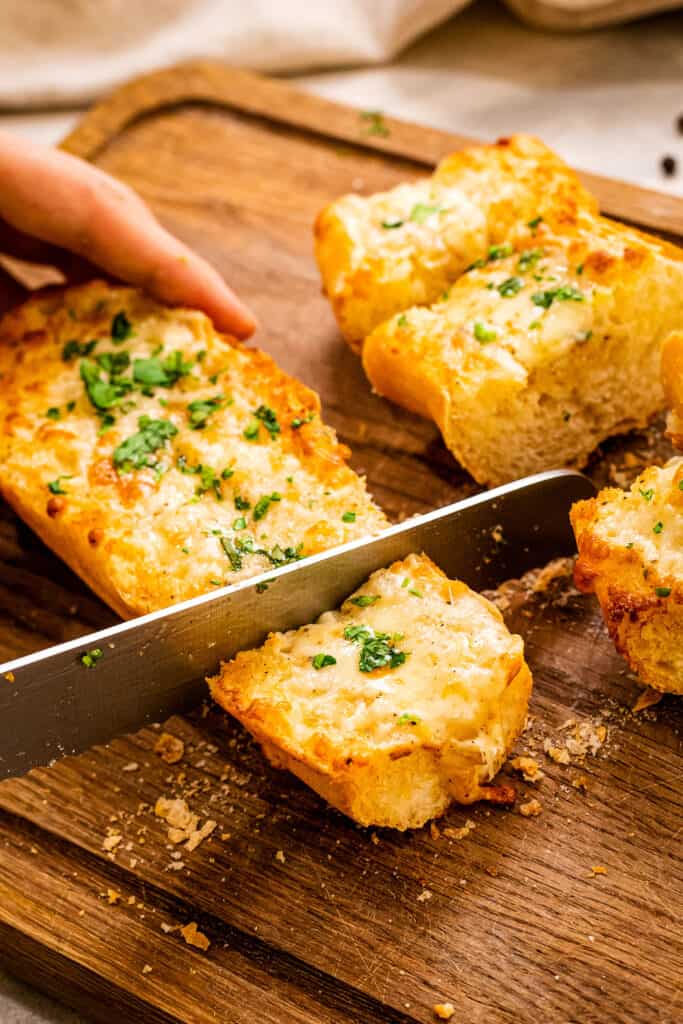 Knife slices cheesy garlic bread on wood cutting board