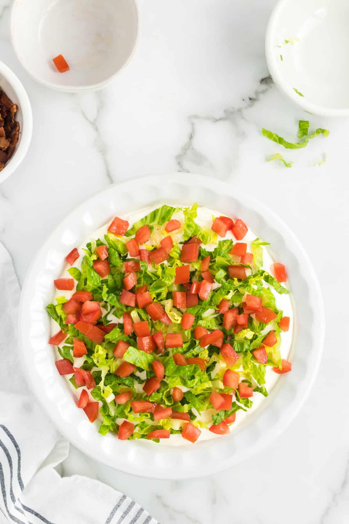 Adding chopped roma tomatos atop lettuce and base layer for BLT Dip in shallow dish