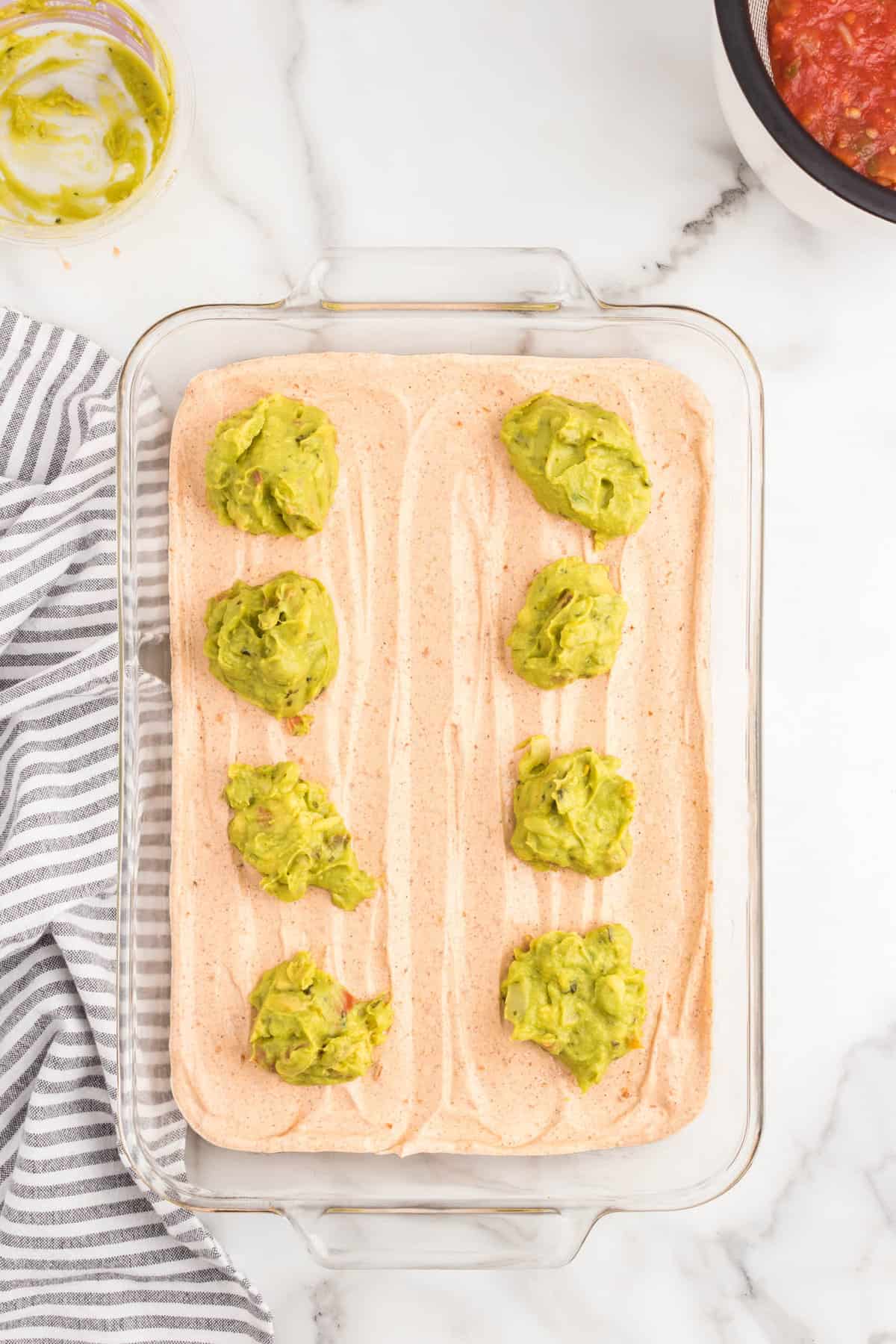 Adding spoonfuls of guacamole atop refried beans in glass serving dish for 7 layer dip