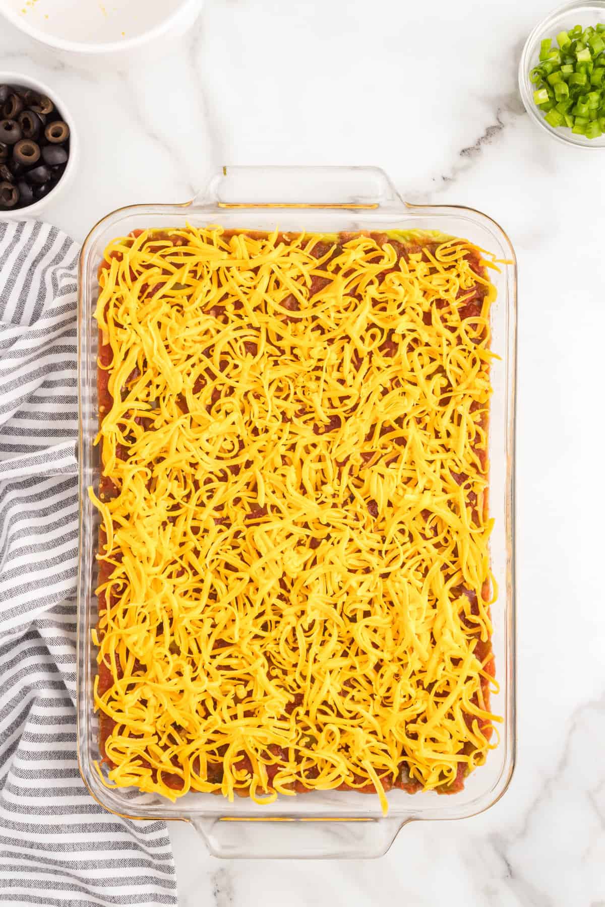 Adding shredded cheddar cheese to 7 layer dip recipe in glass serving dish