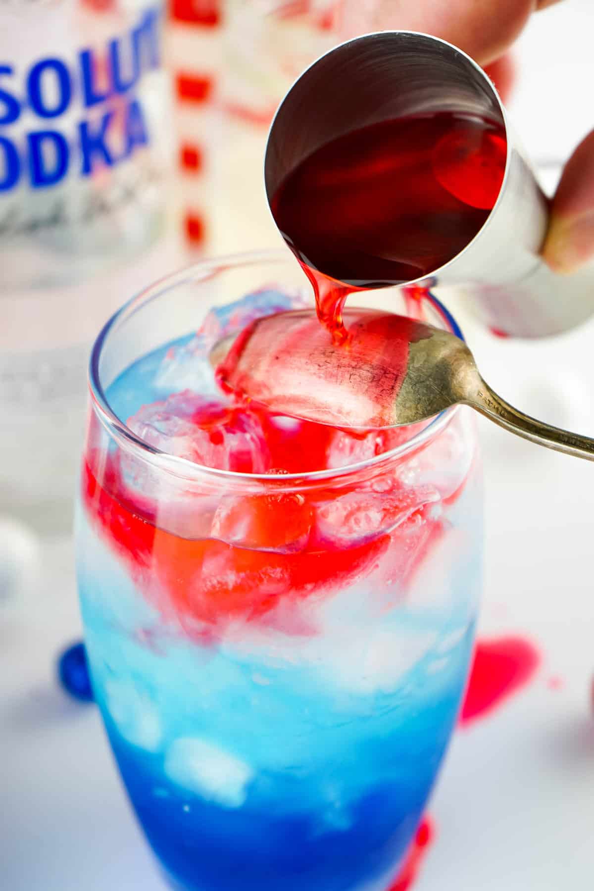 Pouring cherry vodka over spoon to top off Red White and Blue Layered Cocktail 