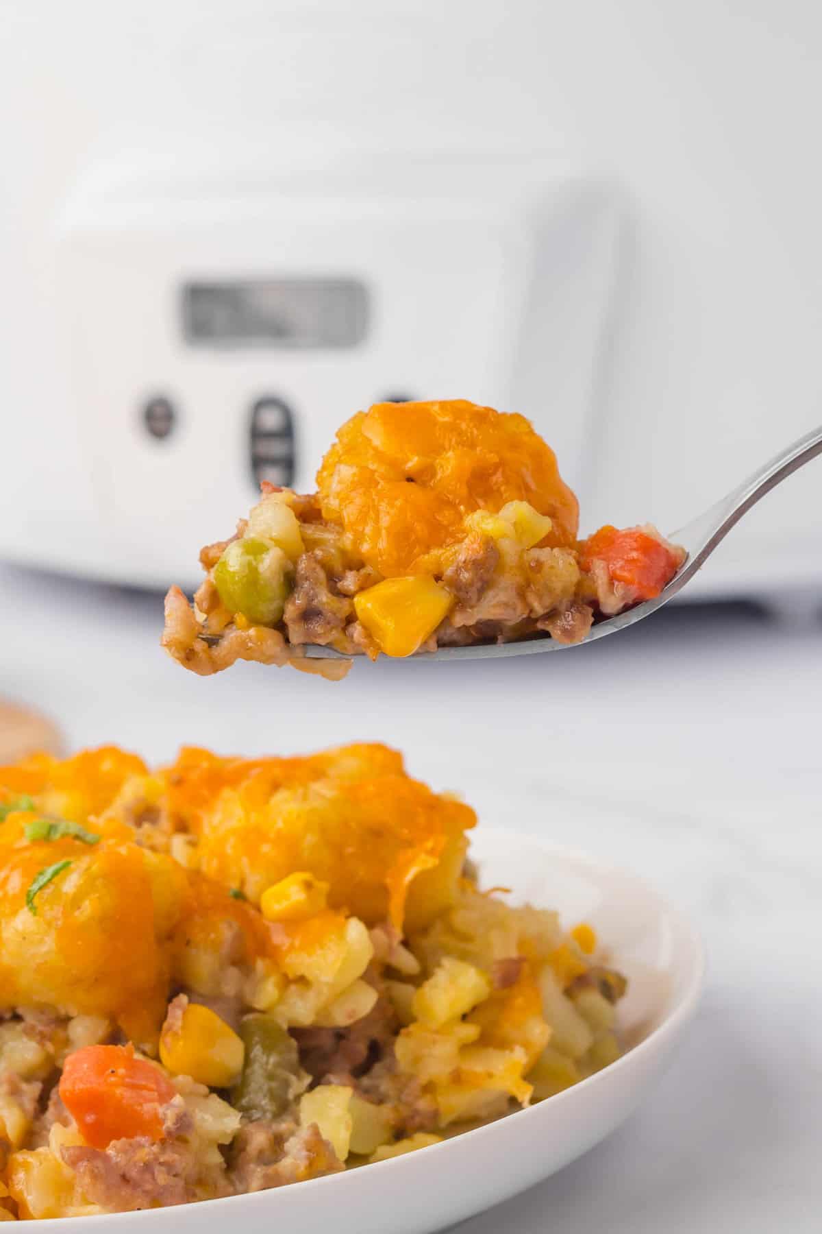 Crock Pot Tater Tot Casserole Recipe on Plate with a Forkful Ready to Enjoy with Crock Pot in Background