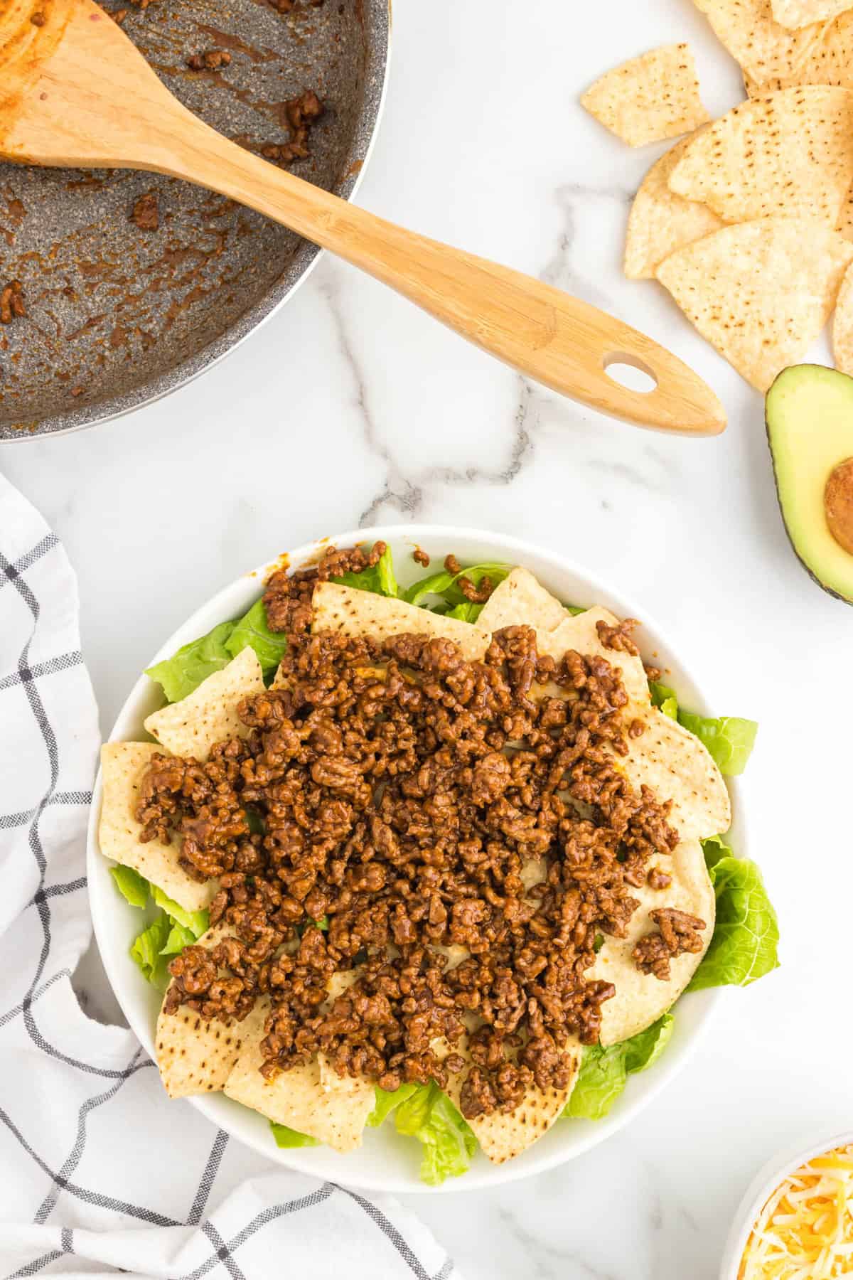 Adding seasoned ground beef to lettuce and tortilla chip layers in bowl for taco salad