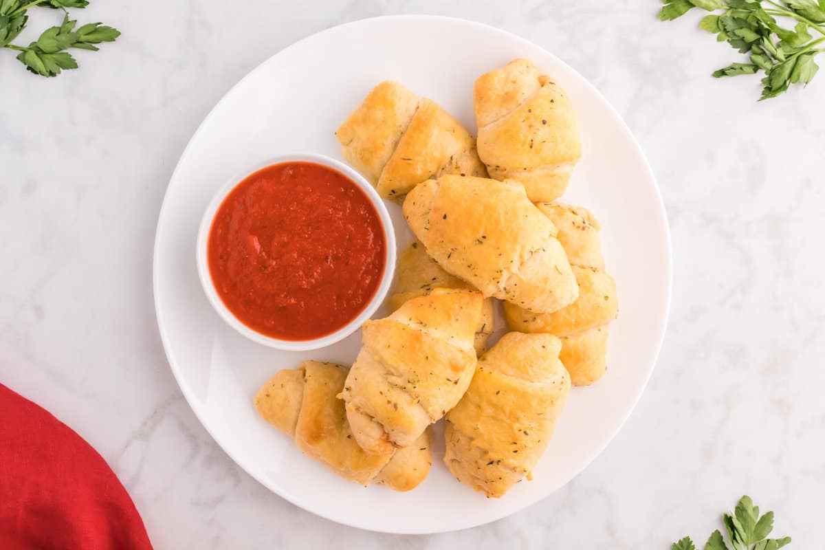 Crescent rolls displayed on a white plate with pizza sauce in a bowl.