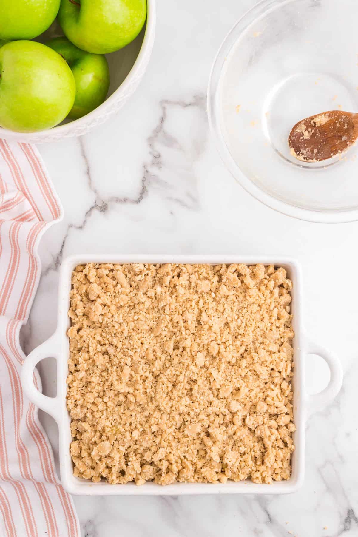 Adding combined topping to seasoned apples in square baking dish for Apple Crumble 