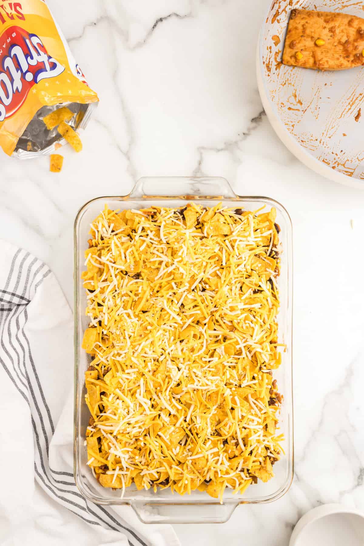 Adding a final layer of shredded cheese to the Frito Pie Casserole in a glass baking dish