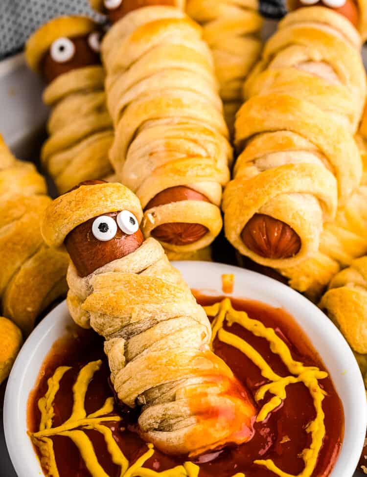 Close up photo of the Mummy Hot Dog sitting inside a white bowl filled with ketchup