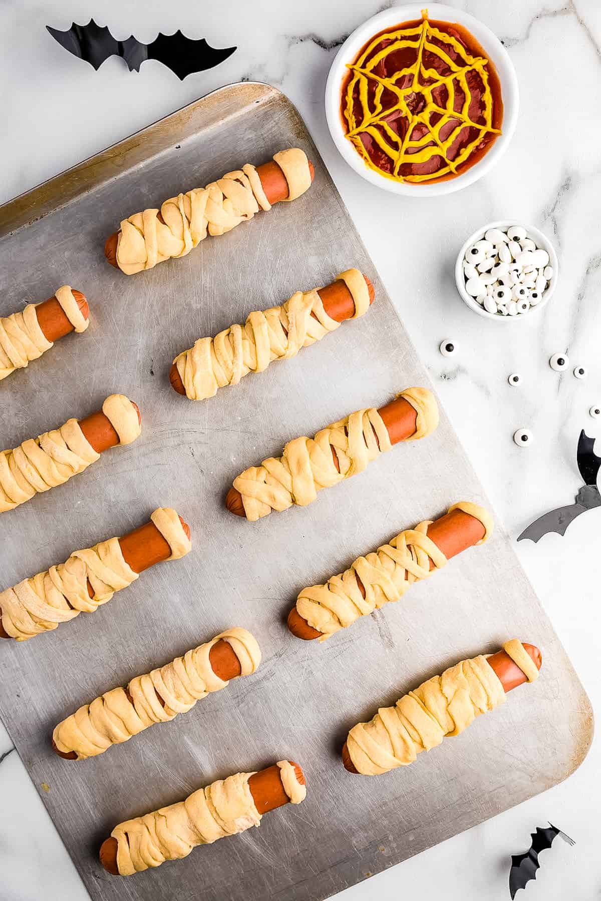 Wrap four Pieces of dough around each hot dog showing portions of the hot dog during the wrapping. Make it look like a Mummy.