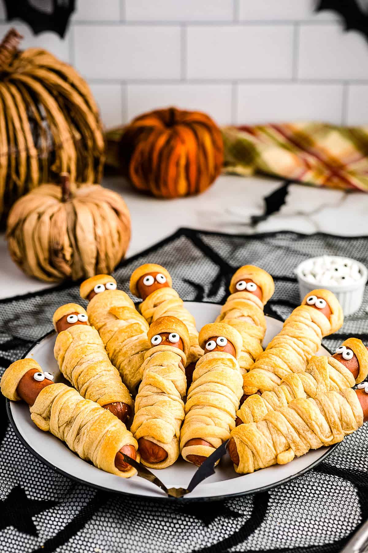 Mummy hot dogs displayed on a white plate with pumpkins in the background.