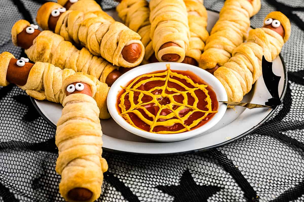 Mummy Hot Dogs displayed on a white plate with a bowl of ketchup and a spider web of mustard.