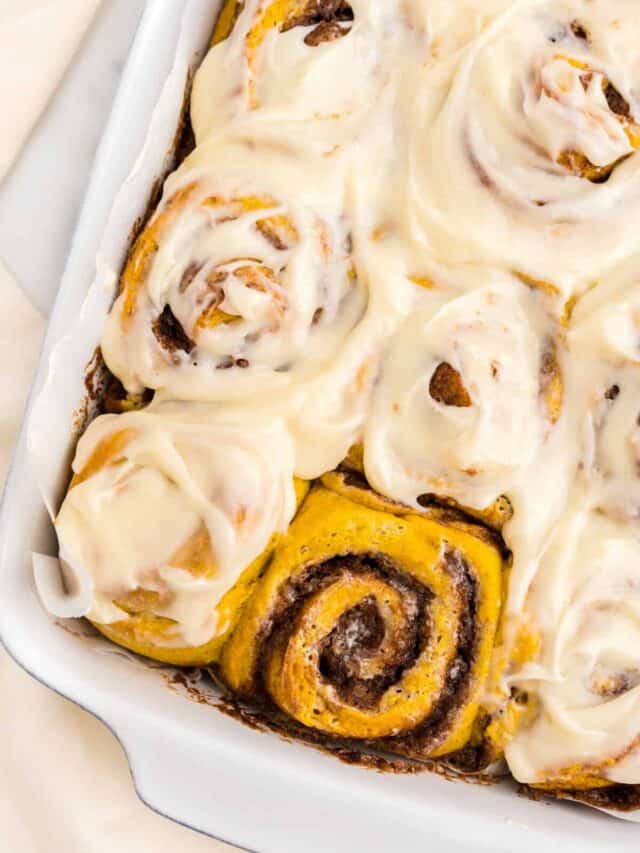 Once cooled, generously spreading cream cheese frosting on Pumpkin Spice Cinnamon Rolls