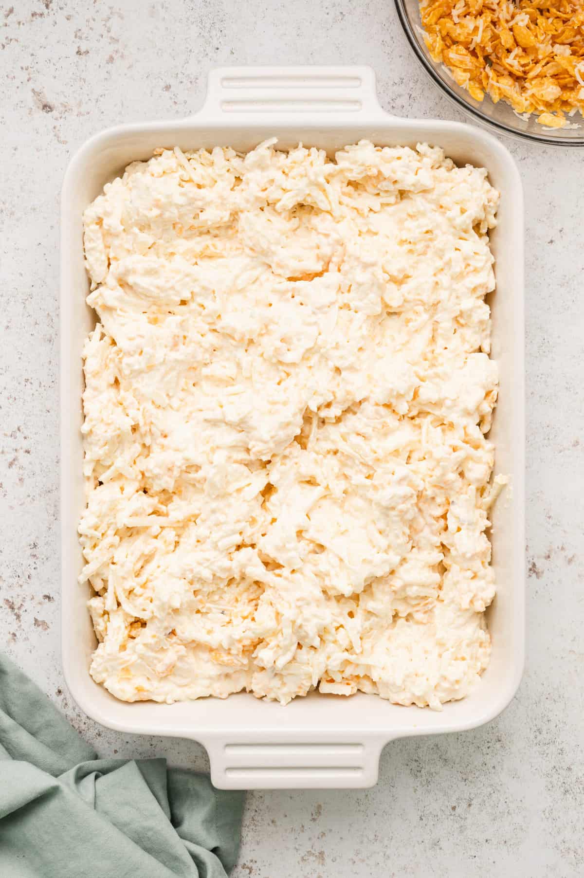 Evenly spreading potatoe mixture in 9x13 baking dish for Cornflakes, melted butter, and Parmesan cheese in glass mixing for Funeral Potatoes recipe