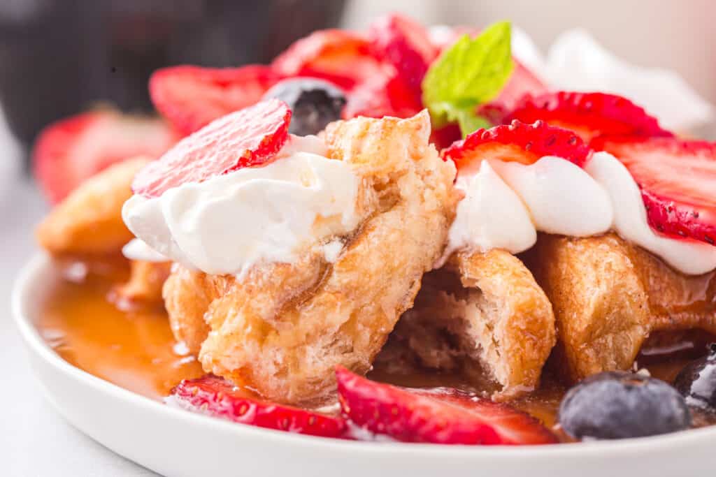 Close up Photo of a Croffle On a white plate with whip cream, fruit and syrup.