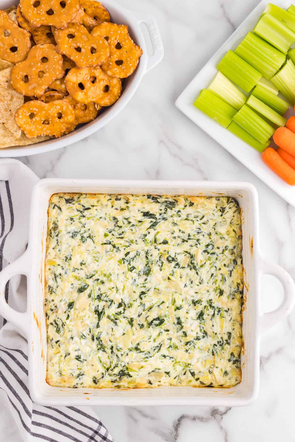 Spinach Artichoke Dip baked to a golden brown in square baking dish with dipper options arranged in bowls & on plates