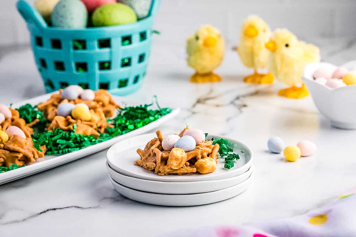 a Stack of little white plates with a Haystack on top. Little Chicks are in the back with a basket full of eggs and another white plate displaying Easter Haystacks.