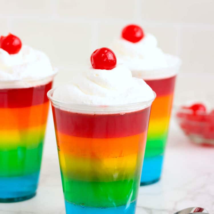 Rainbow Jell-O Cups complete with spoons laying on the table by them.