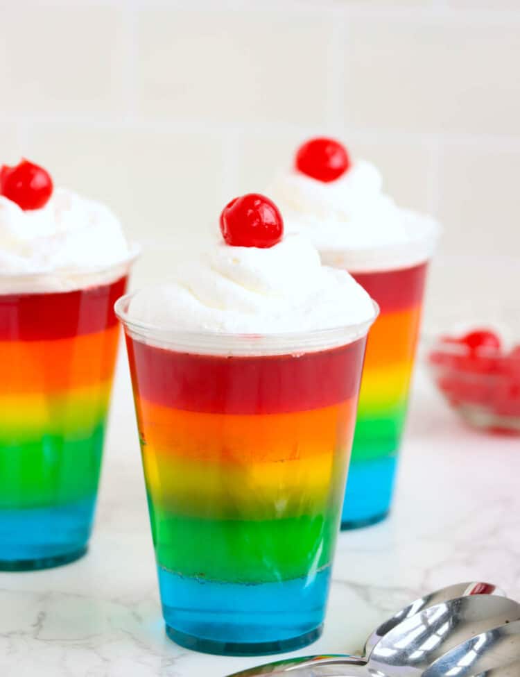 Rainbow Jell-O Cups complete with spoons laying on the table by them.