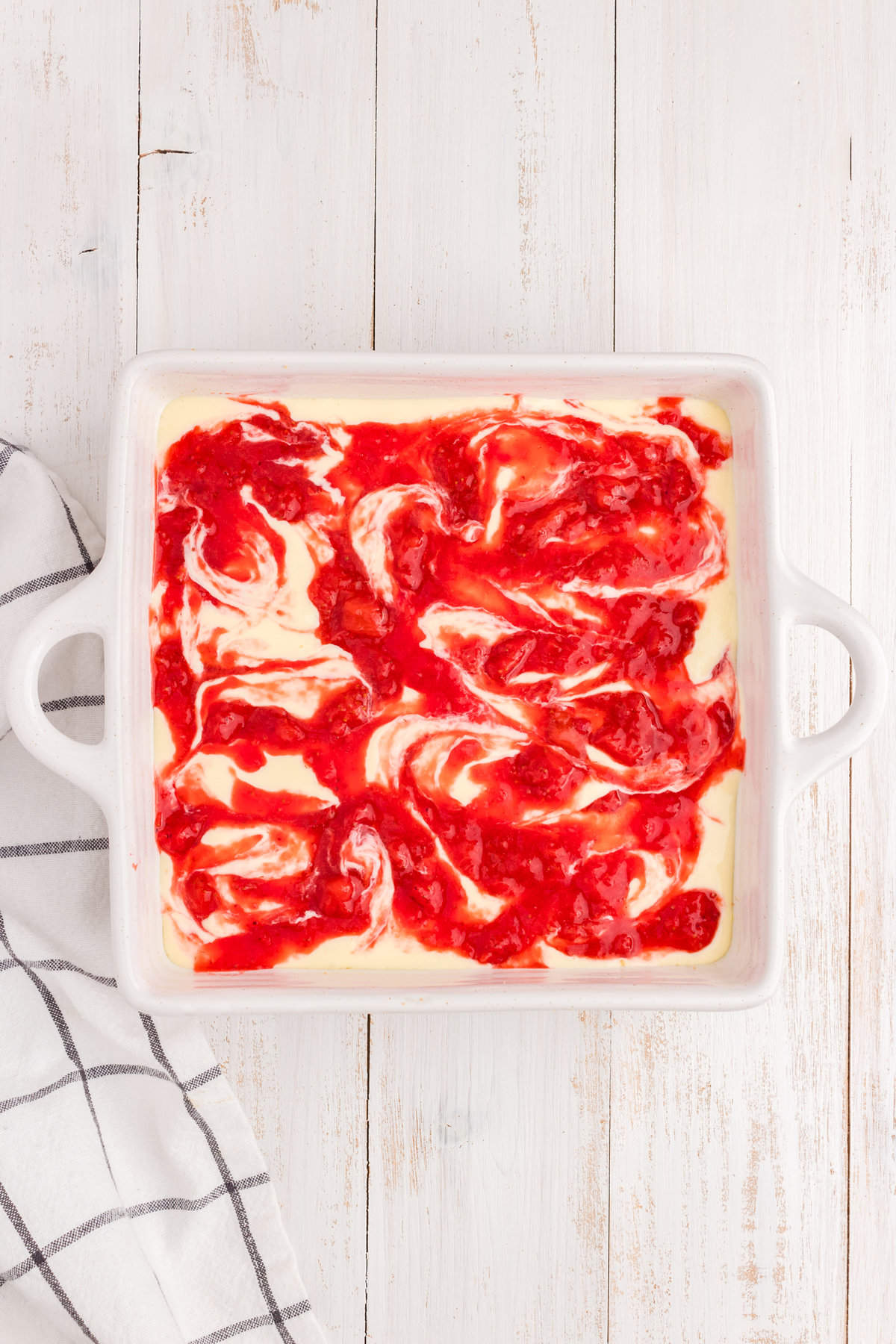 Adding strawberry sauce to cheesecake in square baking dish by swirling for Strawberry Cheesecake Bars recipe