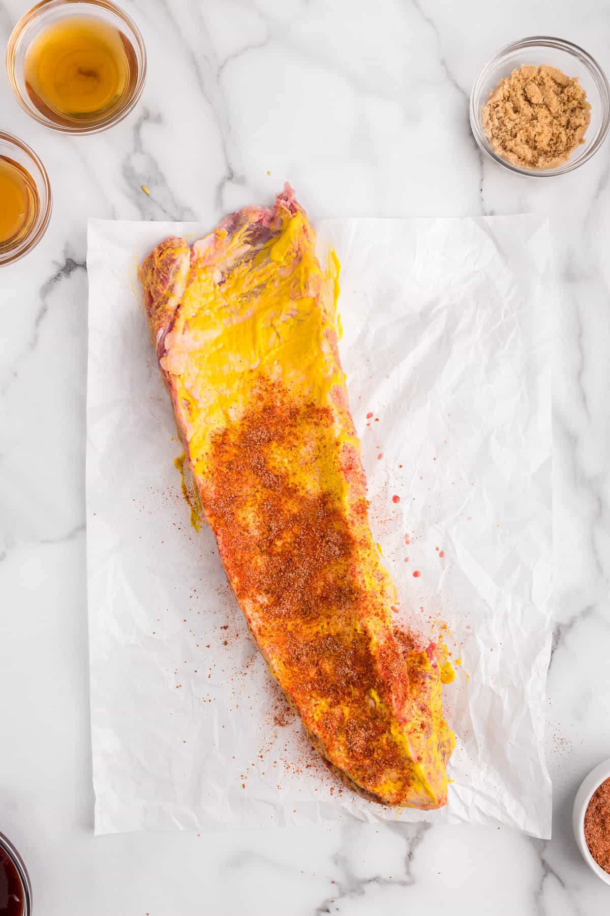 Adding Sweet & Smoky seasoning over mustard on baby back ribs for Brushing mustard on baby back ribs over parchment paper for Oven Baked Ribs recipe recipe