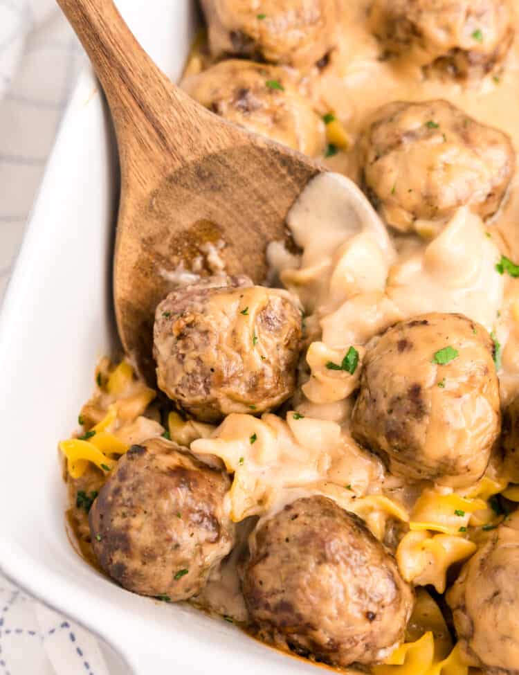 Wooden spoon scooping Swedish Meatball Casserole from white baking dish.