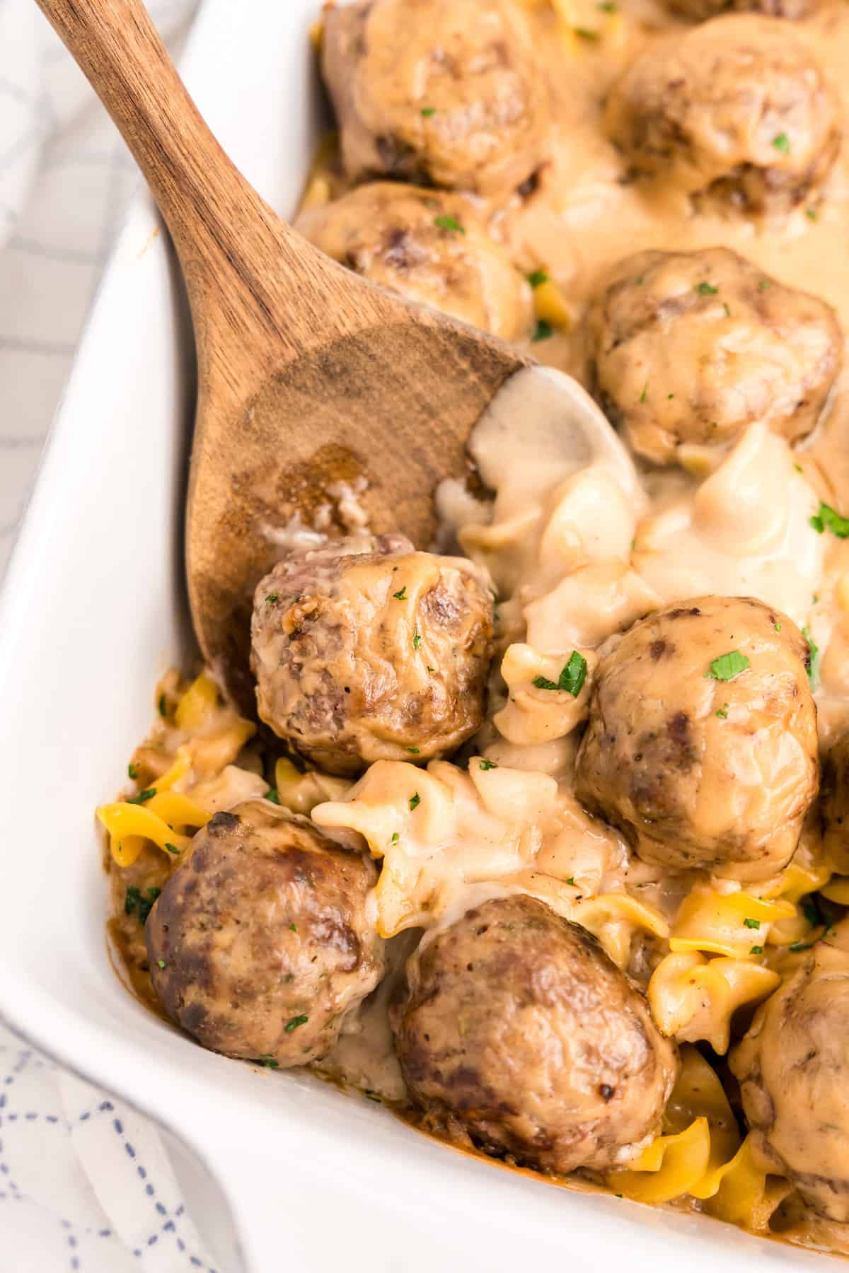 Wooden spoon scooping Swedish Meatball Casserole from white baking dish.